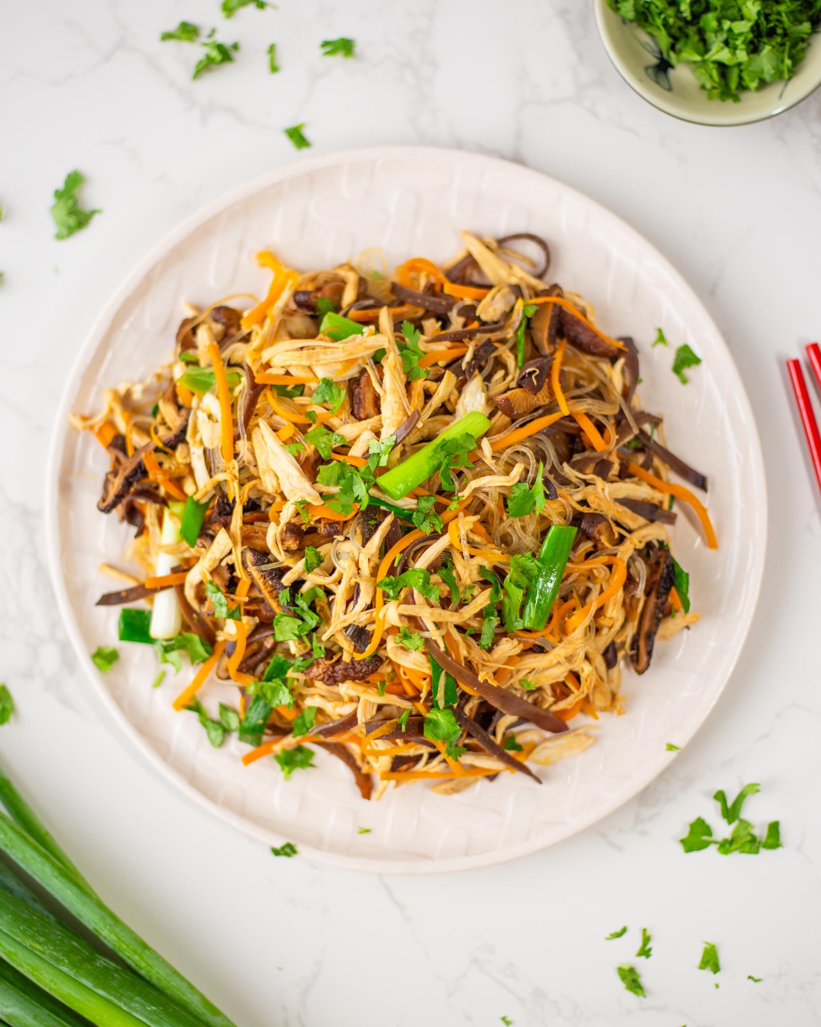 A white plate containing stir-fried Vietnamese glass noodles with shredded chicken, carrot, mushrooms, and green onion.