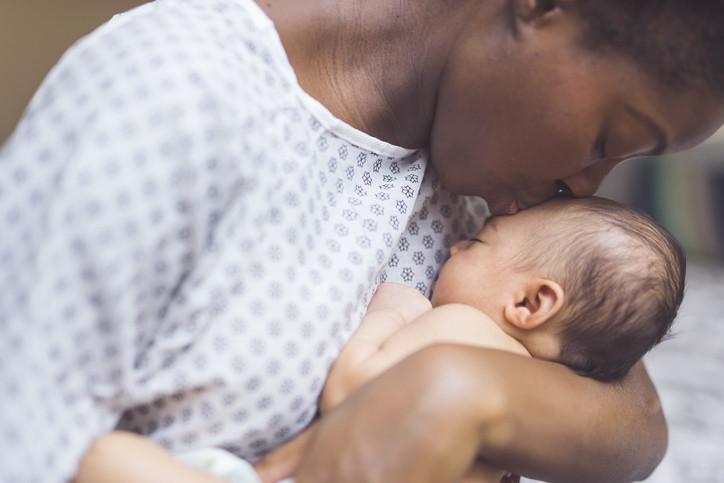 A beautiful young African American mother in a hospital gown gently holds her infant in her arms and smiles down at her. The baby