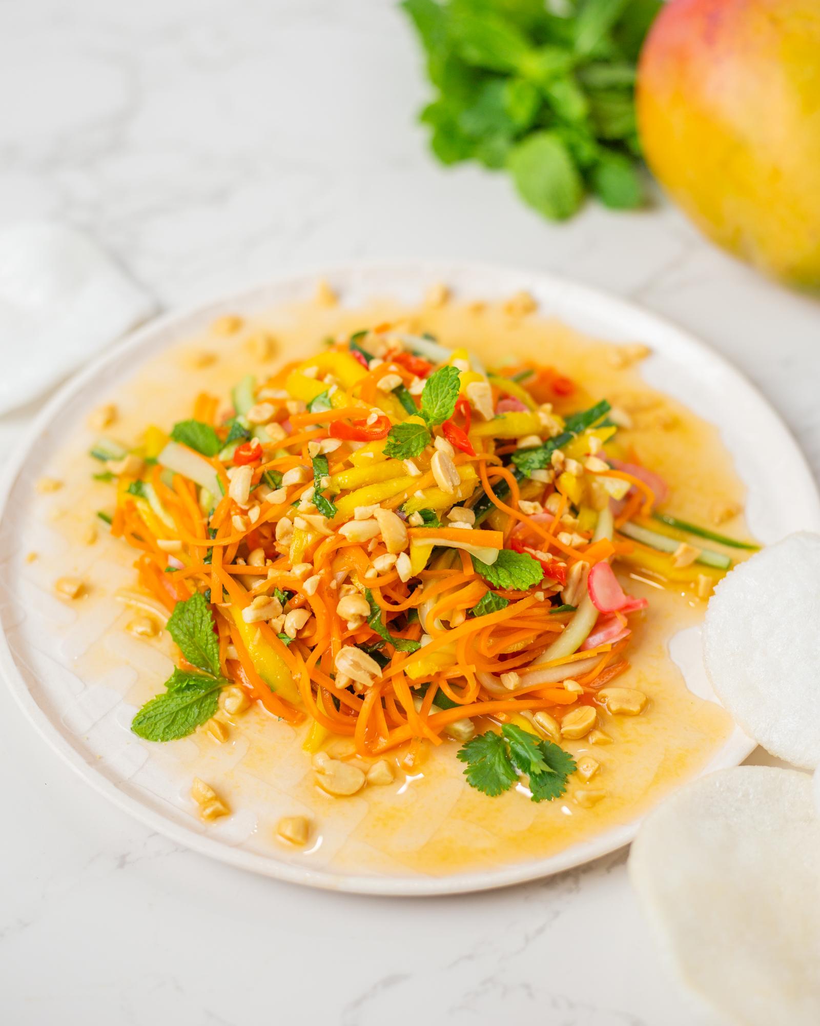 White plate with a colorful salad with strips of mango, carrots, cucumber, and radish; along with herbs and peanuts.