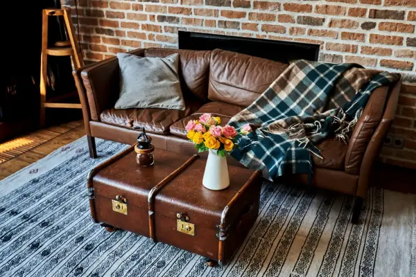 Góc phòng khách với sofa da, gối, chăn, bức tường gạch và vali cũ làm bàn cà phê.
