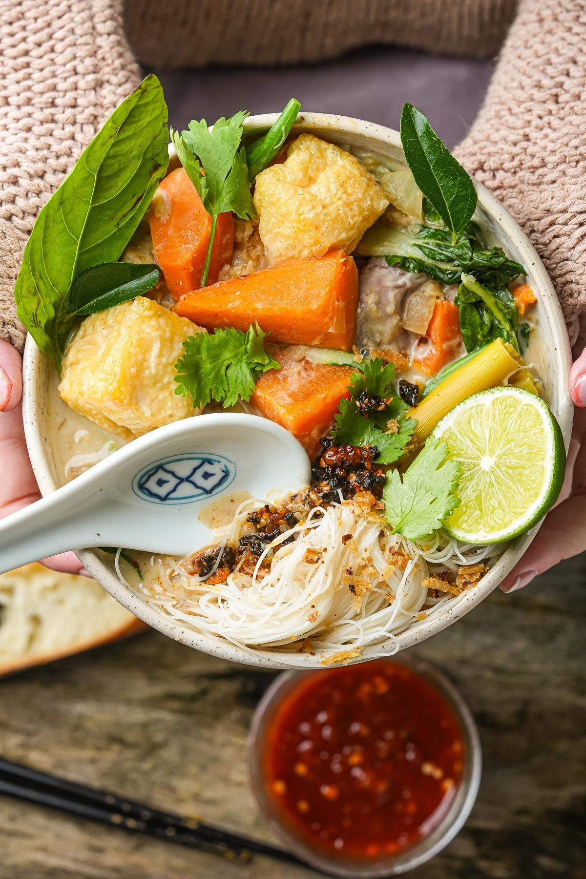 A bowl of cha ri chay with noodles, tofu, carrots, leafy greens, herbs, and a lime wedge being held by a young woman in a sweater.