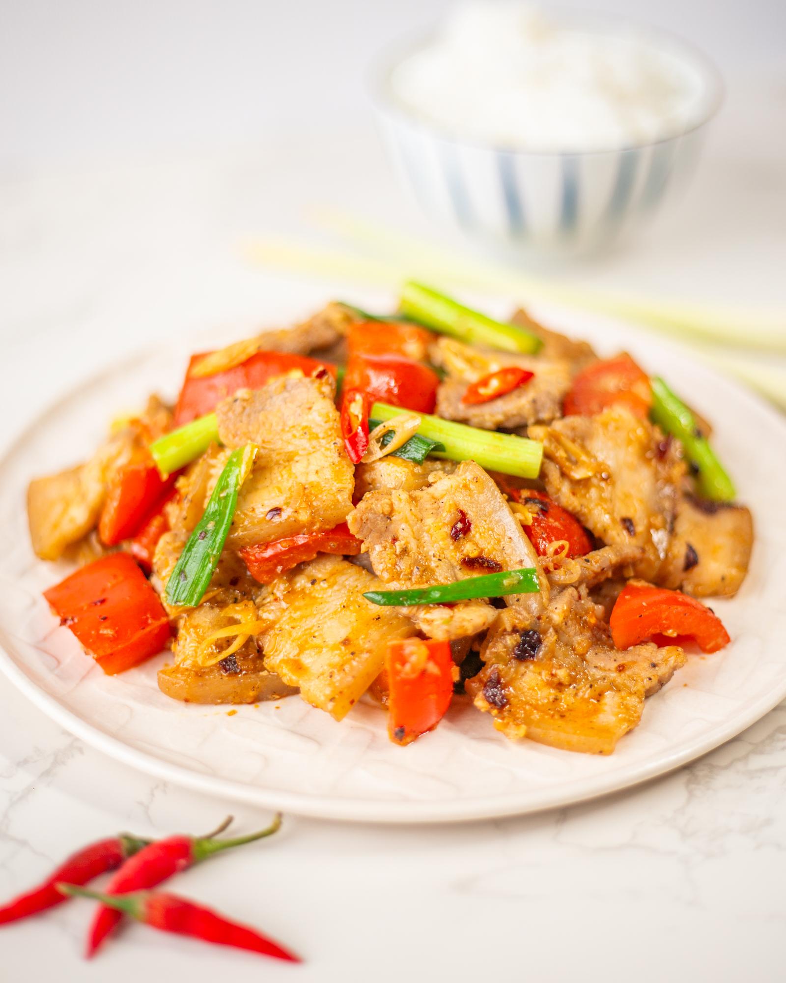 A white plate on a white background. The plate contains thin slices of pork belly that was sautéed with red bell pepper, green onion, lemongrass, chili oil, and other ingredients.