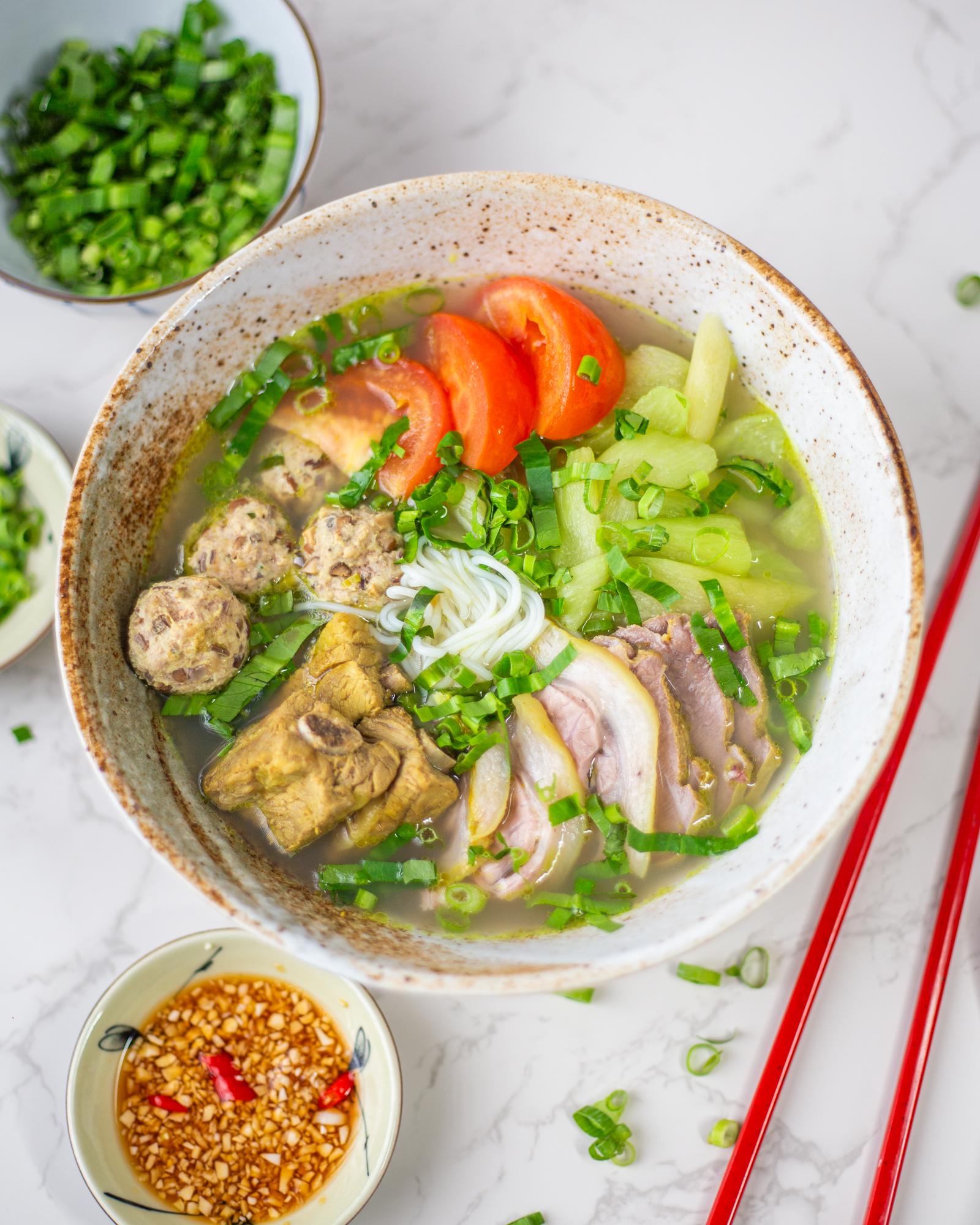 A bowl filled with white rice noodles, different cuts of cooked meat including ribs and meatballs, vegetables, broth, and herbs